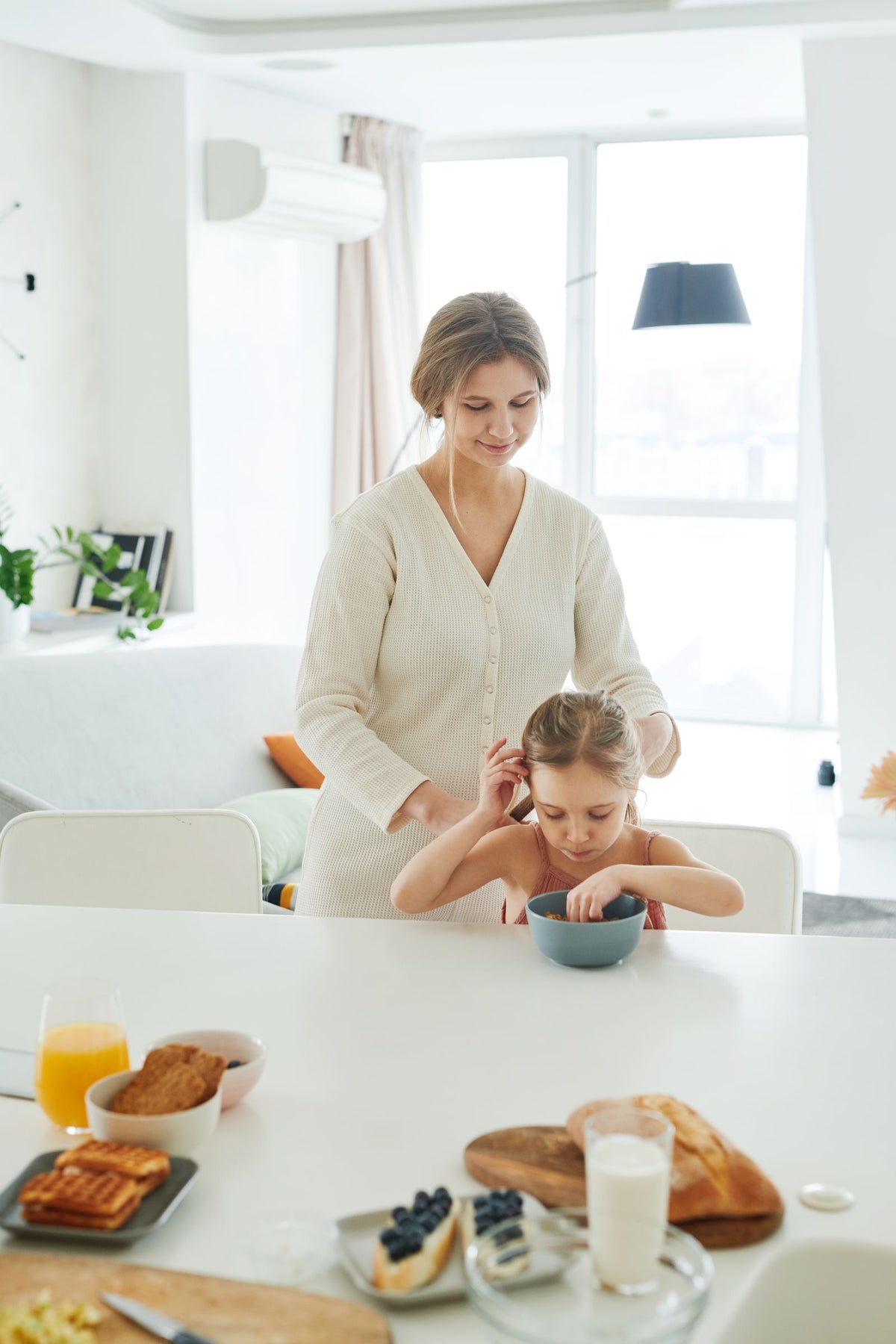 Maman qui attache les cheveux de sa fille dans une cuisine lumineuse au petit-déjeuner Image