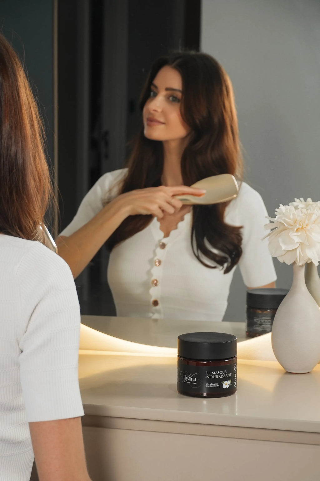Masque Nourrissant à la Phytokératine – Soin Capillaire Luxe Elyara posé sur une table près d’un miroir, flacon noir et personne se coiffant.