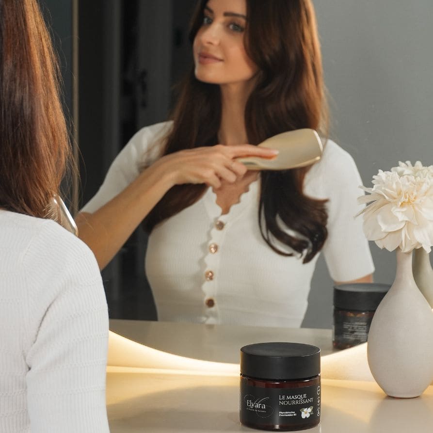 Femme se coiffant devant le miroir avec une brosse; pot noir Nourishing Mask for Dry & Damaged Hair sur la table à côté, vase blanc et fleurs. Image